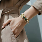 Jakris modern gold cuff bracelet featuring red and green stones worn on a woman's wrist with a beige trench coat.