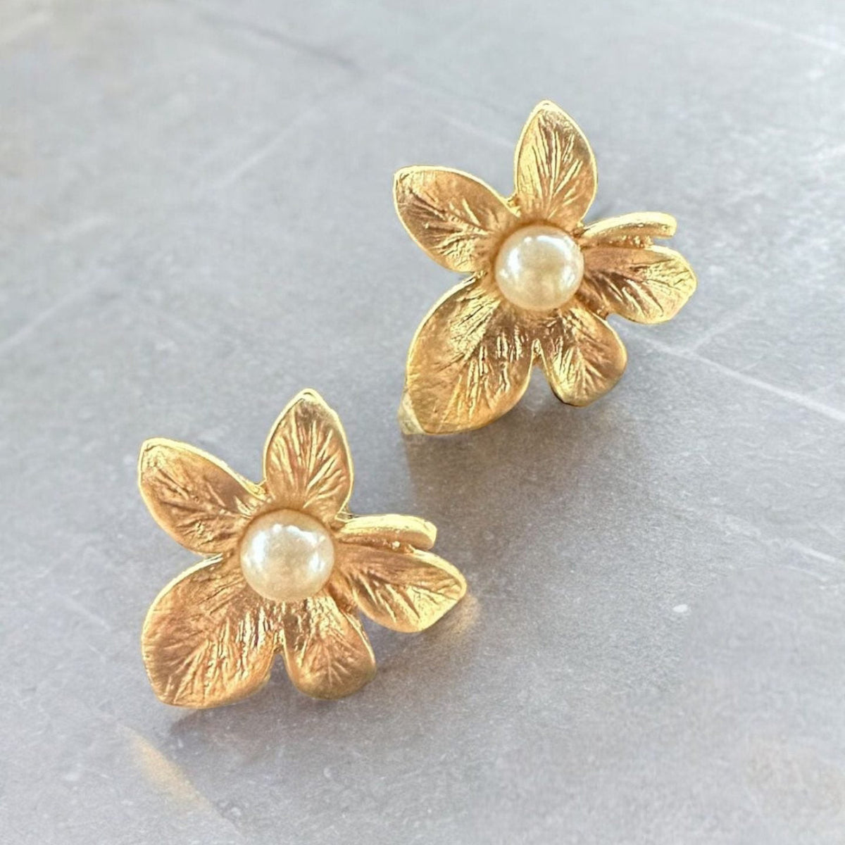 Jakris textured gold-plated flower earrings featuring a central pearl detail on a grey surface