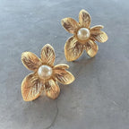 Jakris gold-plated textured flower earrings with central pearl detail on a grey surface