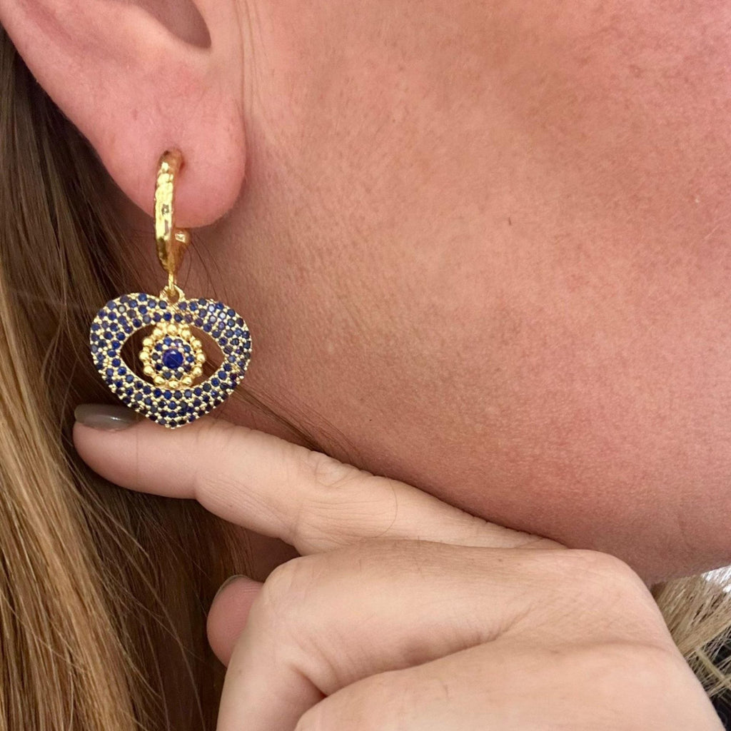 Jakris gold hoop earring featuring a heart-shaped pendant with blue crystal evil eye detailing worn on an earlobe