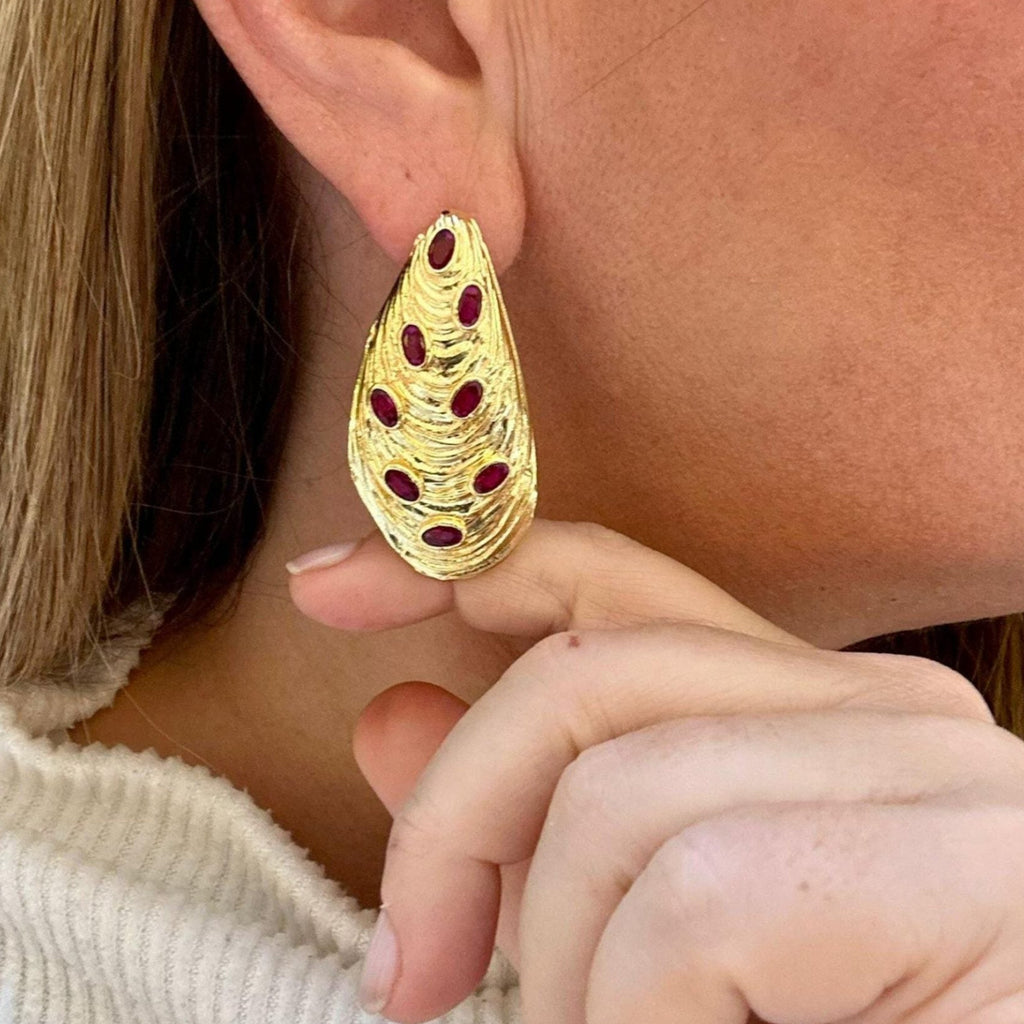 Jakris gold-plated drop earrings featuring a textured organic shape and inlaid red ruby stones worn on an earlobe.