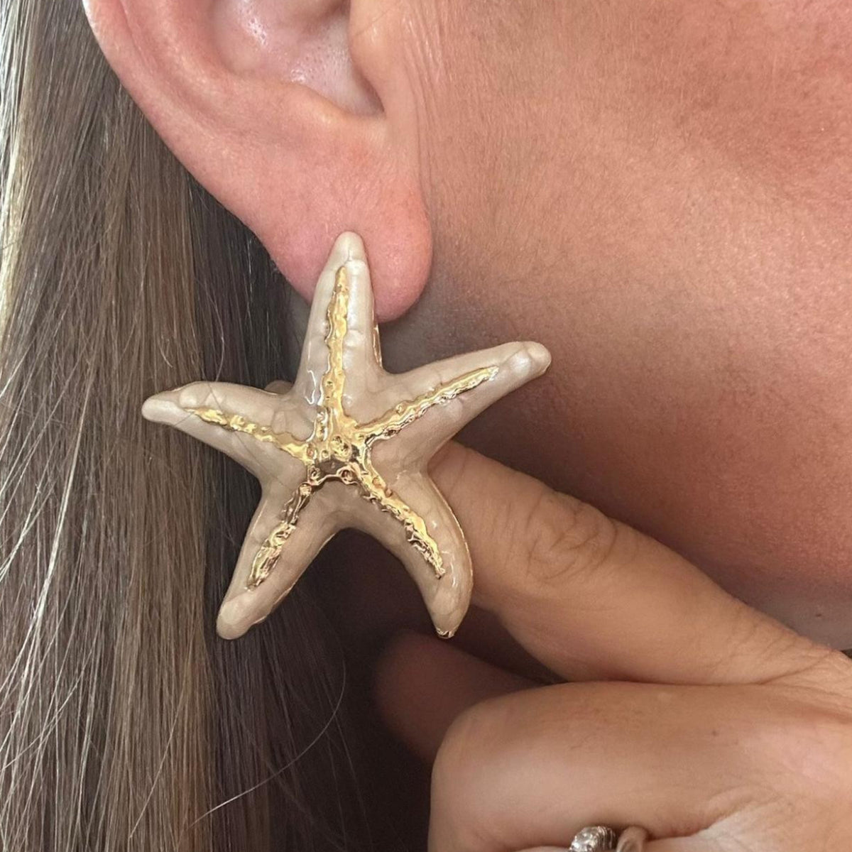 Jakris large starfish enamel earrings in beige and gold worn on a woman's ear for a summer beach style look