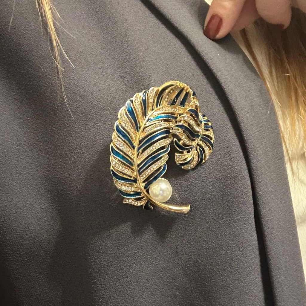 Jakris navy blue enamel feather brooch with gold-tone metal, crystal accents, and a pearl detail on a grey blazer.