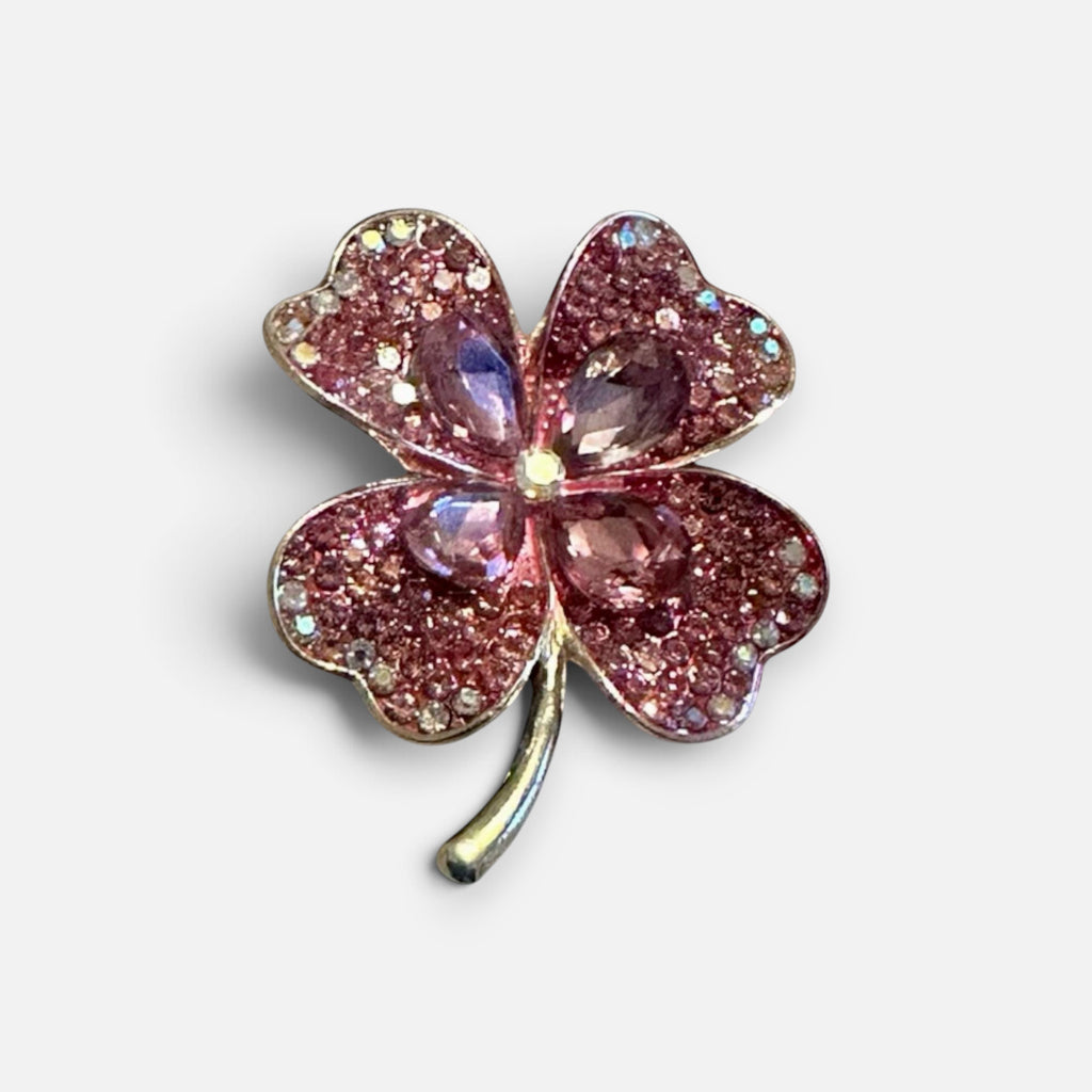 Jakris four-leaf clover brooch featuring sparkling pink crystals and a metallic stem on a white fabric background