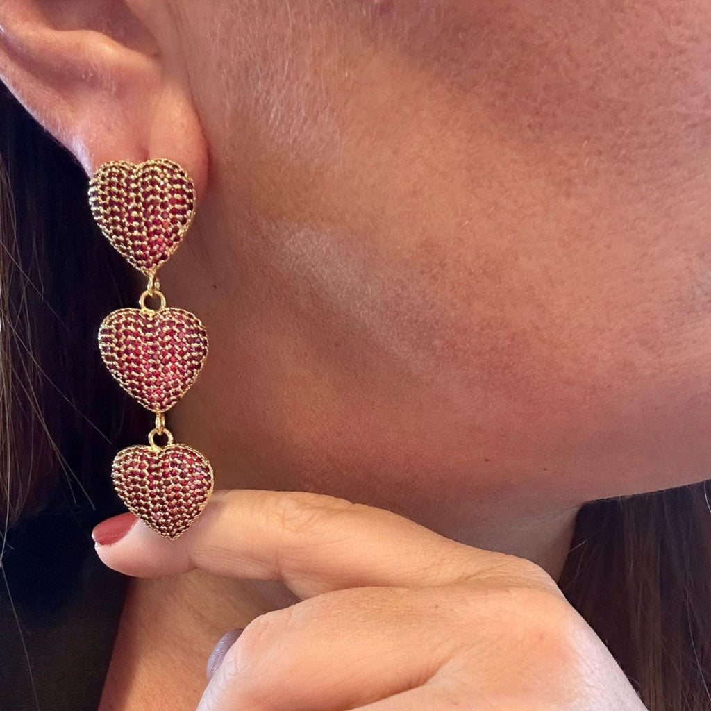 Jakris gold-tone drop earrings featuring three red heart-shaped charms with textured stone detailing on a woman