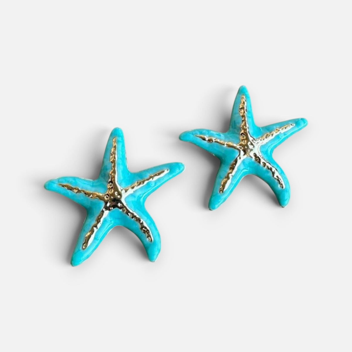 Jakris turquoise and white enamel starfish stud earrings with gold-tone accents on a stone surface