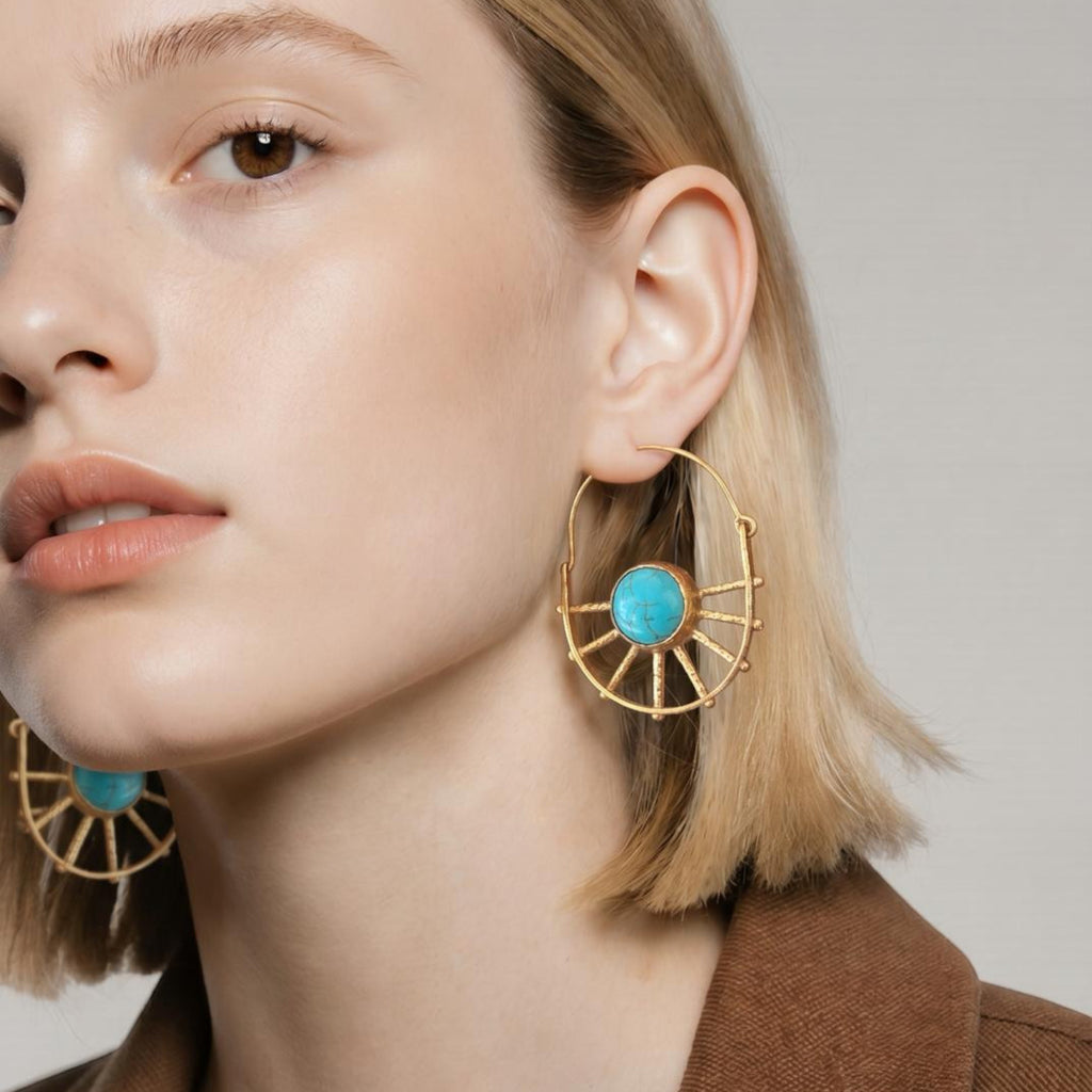 Jakris gold-plated sun-shaped earrings featuring a central turquoise stone worn by a woman with blonde hair.