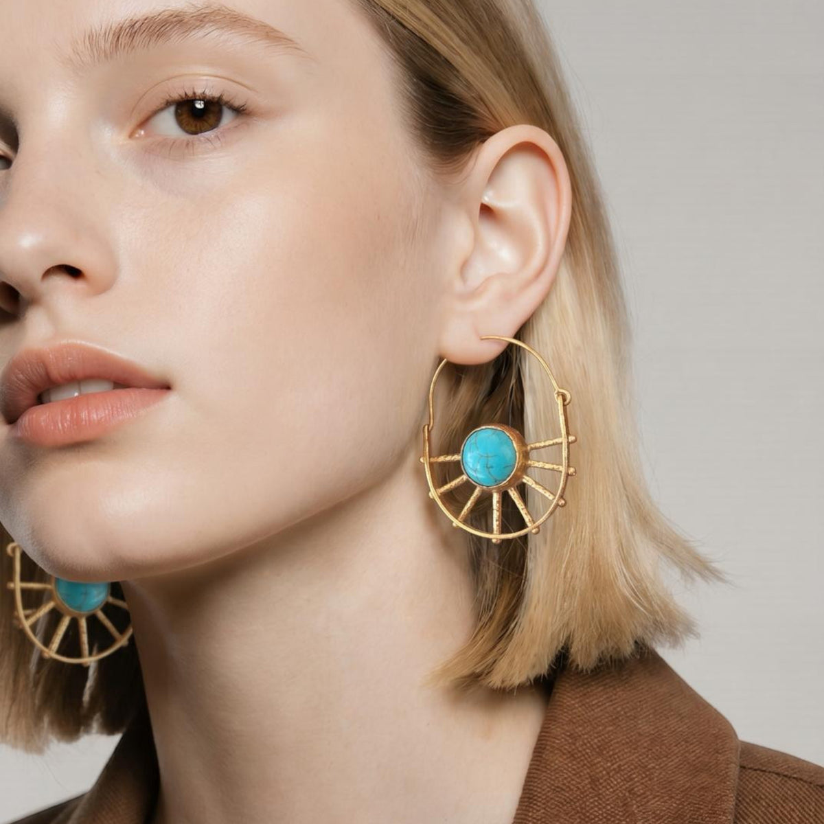 Jakris gold-plated sun-shaped earrings featuring a central turquoise stone worn by a woman with blonde hair.